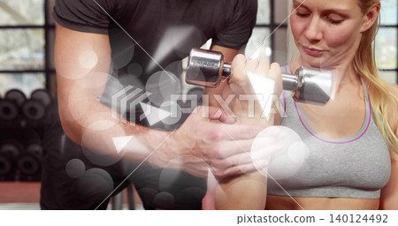 Guiding coach assisting woman in gray sports bra pressing chrome dumbbell in gym his hands spotting Guiding coach assisting woman in gray sports bra pressing chrome dumbbell in gym his hands spotting 140124492