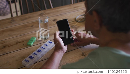 Holding smartphone, senior wearing teal shirt inspecting pill organizer at home table, copy space Holding smartphone, senior wearing teal shirt inspecting pill organizer at home table, copy space 140124533