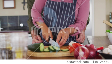 Slicing cucumber, woman in striped apron and partner guiding hands at home kitchen, chef's knife Slicing cucumber, woman in striped apron and partner guiding hands at home kitchen, chef's knife 140124560