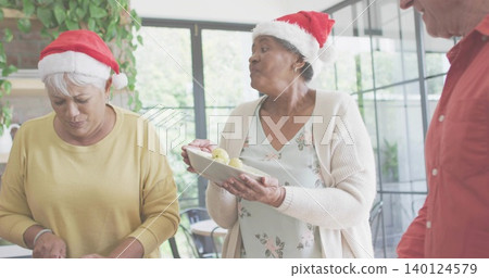 Holding senior woman wearing Santa hat offering white plate with green apples in home kitchen Holding senior woman wearing Santa hat offering white plate with green apples in home kitchen 140124579
