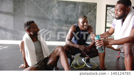 Resting three men wearing athletic wear passing teal water bottle on gym floor, with jump rope Resting three men wearing athletic wear passing teal water bottle on gym floor, with jump rope 140124785
