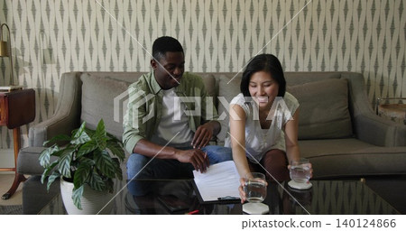 Leaning couple on sofa in casual clothing reviewing clipboard papers at home, with glass tumblers Leaning couple on sofa in casual clothing reviewing clipboard papers at home, with glass tumblers 140124866