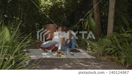 Sitting couple wearing white blouse and light green overshirt on picnic blanket in garden, with urn 140124867