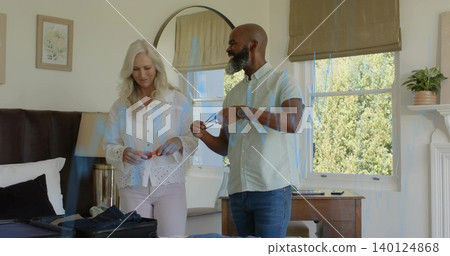 Exchanging eyeglasses, man in jeans and senior woman wearing blouse by bed with open suitcase 140124868
