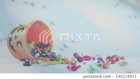 Tipping orange jack-o'-lantern bucket spilling foil candies across pale tabletop, confetti backdrop Tipping orange jack-o'-lantern bucket spilling foil candies across pale tabletop, confetti backdrop 140124921