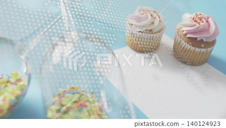 Showing two sprinkled cupcakes on pastel-blue table, inverted glasses, dotted light, copy space 140124923