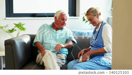 Checking provider in blue scrubs measuring blood pressure for senior man on clinic sofa with cuff Checking provider in blue scrubs measuring blood pressure for senior man on clinic sofa with cuff 140125103