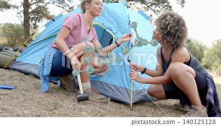 Kneeling women erecting blue dome tent in camp clearing, using mallet, poles, flannel waists, boots Kneeling women erecting blue dome tent in camp clearing, using mallet, poles, flannel waists, boots 140125128