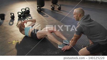 Woman trainee doing sit-ups in sports bra and shorts, coach holding feet on gym floor, kettlebells Woman trainee doing sit-ups in sports bra and shorts, coach holding feet on gym floor, kettlebells 140125220
