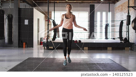 Skipping woman in athletic wear using jump-rope on mat in industrial gym with boxing ring, bags Skipping woman in athletic wear using jump-rope on mat in industrial gym with boxing ring, bags 140125523