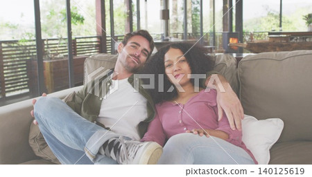 Sitting couple wearing casual clothes cuddling on light-beige sofa in home lounge, with glass doors Sitting couple wearing casual clothes cuddling on light-beige sofa in home lounge, with glass doors 140125619