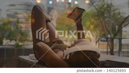 Reclining woman in dark athleisure holding smartphone overhead on concrete ledge, white headphones Reclining woman in dark athleisure holding smartphone overhead on concrete ledge, white headphones 140125693