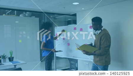 Placing yellow sticky note, woman holding tablet in blue top collaborating with man at whiteboard Placing yellow sticky note, woman holding tablet in blue top collaborating with man at whiteboard 140125925