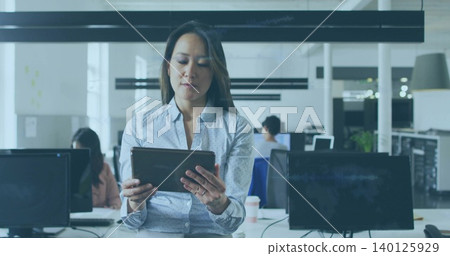 Holding tablet, woman in light blue patterned shirt standing at open-plan office desk with monitors Holding tablet, woman in light blue patterned shirt standing at open-plan office desk with monitors 140125929