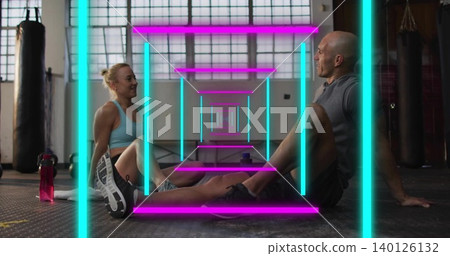 Sitting gym partners in activewear facing each other on rubber gym floor, punching bags, neon bars Sitting gym partners in activewear facing each other on rubber gym floor, punching bags, neon bars 140126132
