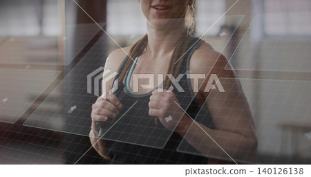 Holding jump rope, midadult woman in blacktank and blue-sports-bra posing in gym with window grid Holding jump rope, midadult woman in blacktank and blue-sports-bra posing in gym with window grid 140126138