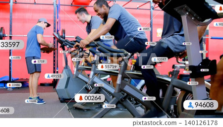 Pedaling man wearing navy t-shirt and dark shorts on spin bike at fitness studio, with badges 140126178