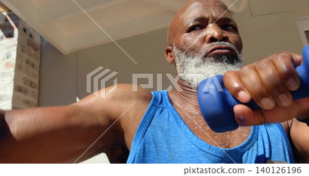 Lifting gray-bearded man holding blue dumbbell on covered porch, wearing bright blue tank top Lifting gray-bearded man holding blue dumbbell on covered porch, wearing bright blue tank top 140126196