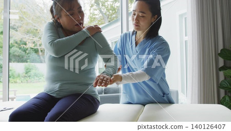 Guiding clinician in scrubs supporting patient extending arm on padded table, sliding glass doors Guiding clinician in scrubs supporting patient extending arm on padded table, sliding glass doors 140126407
