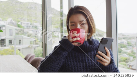 Sipping woman wearing navy sweater, holding red mug and checking smartphone at home by windows Sipping woman wearing navy sweater, holding red mug and checking smartphone at home by windows 140126929