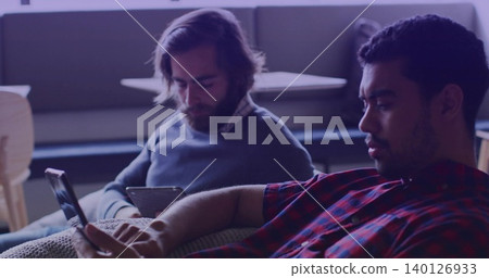 Sitting two men on couch in lounge, wearing red plaid, gray pullover, using smartphone and tablet Sitting two men on couch in lounge, wearing red plaid, gray pullover, using smartphone and tablet 140126933
