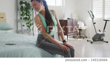 Sitting Korean woman wearing mint top and gray leggings in sunny bedroom, fitness tracker and phone Sitting Korean woman wearing mint top and gray leggings in sunny bedroom, fitness tracker and phone 140126988