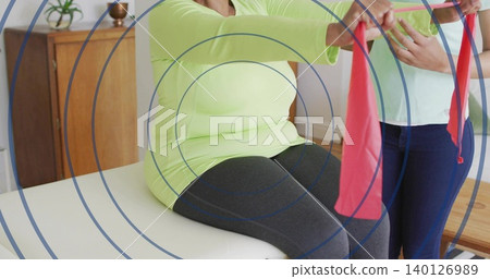 Helping therapist guiding senior woman in lime-top doing band work on clinic table, target overlay Helping therapist guiding senior woman in lime-top doing band work on clinic table, target overlay 140126989