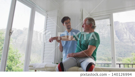 Guiding therapist supporting man in green tee on padded table by windows, knee supports, copy space Guiding therapist supporting man in green tee on padded table by windows, knee supports, copy space 140126994