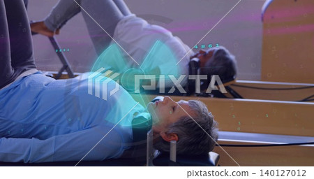 Lying senior woman performing reformer Pilates at fitness studio wearing blue top with teal overlay 140127012