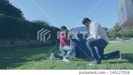 Kneeling mature man and boy inspecting vertical solar panel on backyard lawn, showing wristwatch Kneeling mature man and boy inspecting vertical solar panel on backyard lawn, showing wristwatch 140127056