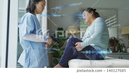 Standing nurse in blue scrubs observing seated patient holding knee on ottoman at home, waveform Standing nurse in blue scrubs observing seated patient holding knee on ottoman at home, waveform 140127085