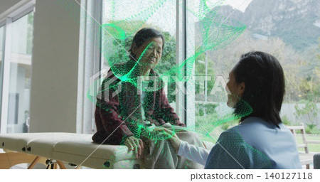 Holding hands caregiver in light-blue scrubs kneeling at table by glass doors in home, green mesh Holding hands caregiver in light-blue scrubs kneeling at table by glass doors in home, green mesh 140127118