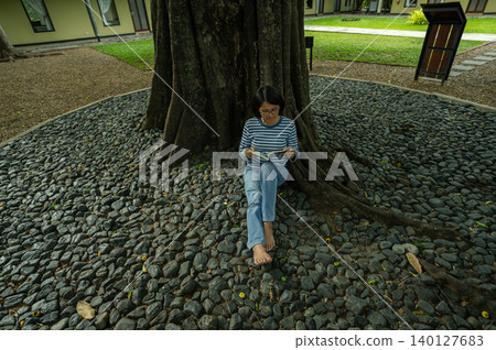 Thai woman sitting barefoot reading book under tree at luxury hotel resort. Peaceful vacation retreat. Digital detox and mindful wellness relaxation. Quiet reading time under garden tree at hotel. Thai woman sitting barefoot reading book under tree at luxury hotel resort. Peaceful vacation retreat. Digital detox and mindful wellness relaxation. Quiet reading time under garden tree at hotel. 140127683