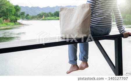 A woman with a cloth bag sitting on a wooden riverfront deck gazing at the calm river, symbolizing slow living, digital detox, solo retreat, and emotional recharge in nature for self care and balance. 140127707