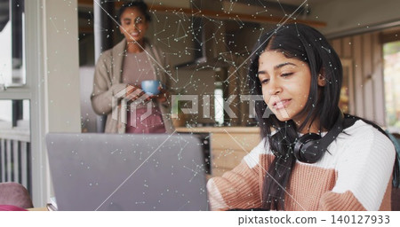 Typing teen girl in sweater headset using laptop at kitchen table mother holding blue mug watching Typing teen girl in sweater headset using laptop at kitchen table mother holding blue mug watching 140127933