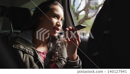 Talking adult woman in green jacket holding smartphone to mouth in back seat, window light Talking adult woman in green jacket holding smartphone to mouth in back seat, window light 140127941
