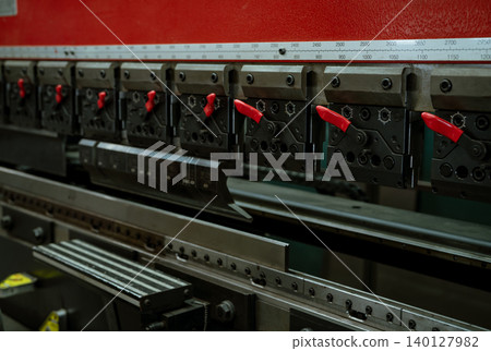 CNC metal press brake with aligned tooling and red locking handles. Operators shape steel components in an industrial workshop. Precision, controlled force and accurate setup every fabrication step. 140127982