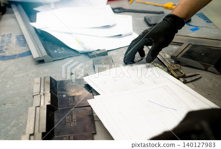 Worker's hand in black gloves examining technical drawings and metal tools on a workshop table, highlighting the precision and skill involved in industrial craftsmanship and manufacturing planning. Worker's hand in black gloves examining technical drawings and metal tools on a workshop table, highlighting the precision and skill involved in industrial craftsmanship and manufacturing planning. 140127983