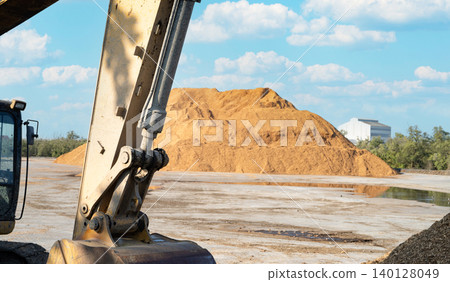 Excavator bucket and hydraulic arm at industrial yard with large sawdust pile, showing heavy machinery handling biomass material for wood processing, recycling, and sustainable industrial production. Excavator bucket and hydraulic arm at industrial yard with large sawdust pile, showing heavy machinery handling biomass material for wood processing, recycling, and sustainable industrial production. 140128049