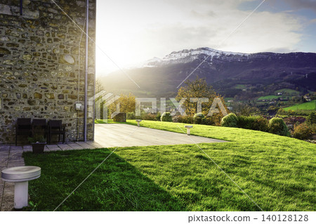 Sunlit countryside landscape with green meadow, stone house terrace, and mountain valley in autumn. Peaceful rural living, natural environment, and travel destination for relaxation and wellbeing. 140128128