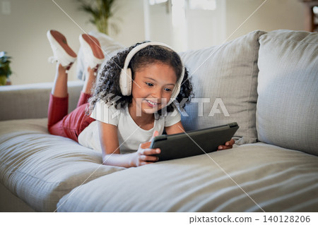 African American girl lying on couch in living room using tablet wearing headphones near plant African American girl lying on couch in living room using tablet wearing headphones near plant 140128206