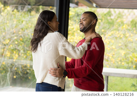 Diverse couple embracing on modern veranda through glass wall by railing overlooking yellow trees Diverse couple embracing on modern veranda through glass wall by railing overlooking yellow trees 140128216