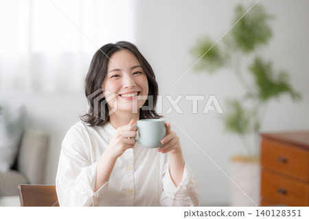 一位微笑的女士手捧馬克杯,展現出輕鬆的晨間生活狀態,上半身面向鏡頭。 一位微笑的女士手捧馬克杯,展現出輕鬆的晨間生活狀態,上半身面向鏡頭。 140128351