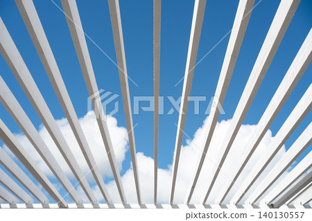 Symmetrical low angle view of white wooden pergola beams converging against a blue cloudy sky. Abstract architectural background Symmetrical low angle view of white wooden pergola beams converging against a blue cloudy sky. Abstract architectural background 140130557