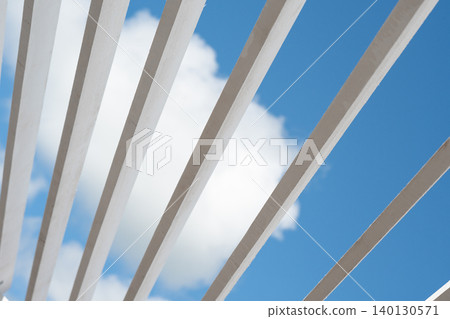 Minimalist geometric pattern of a white sunshade structure against a bright blue sky with a cloud. Abstract architectural background with white wooden pergola planks and a fluffy cloud in the blue Minimalist geometric pattern of a white sunshade structure against a bright blue sky with a cloud. Abstract architectural background with white wooden pergola planks and a fluffy cloud in the blue 140130571
