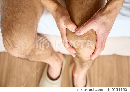 Man suffering from knee pain, gripping and massaging bare knee with both hands while sitting on bed, top view. Concept of joint discomfort and health care. Man suffering from knee pain, gripping and massaging bare knee with both hands while sitting on bed, top view. Concept of joint discomfort and health care. 140131160