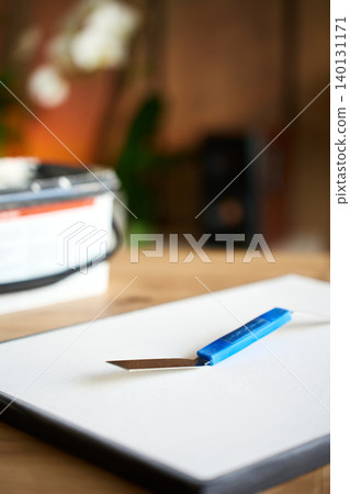 A blue palette knife rests on white canvas beside a paste container on a wooden table. Concept of DIY textured artwork and craft preparation. A blue palette knife rests on white canvas beside a paste container on a wooden table. Concept of DIY textured artwork and craft preparation. 140131171
