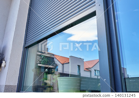 Grey exterior roller shutter is partially open above a large glass window on a modern house facade, with sky, neighboring houses and indoor plants visible through the glass. 140131245
