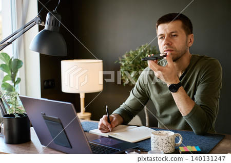 Man speaking to smartphone and writing notes at home desk with laptop, coffee mug and warm interior light. Concept of voice assistant, daily planning, remote work and everyday AI technology. Man speaking to smartphone and writing notes at home desk with laptop, coffee mug and warm interior light. Concept of voice assistant, daily planning, remote work and everyday AI technology. 140131247