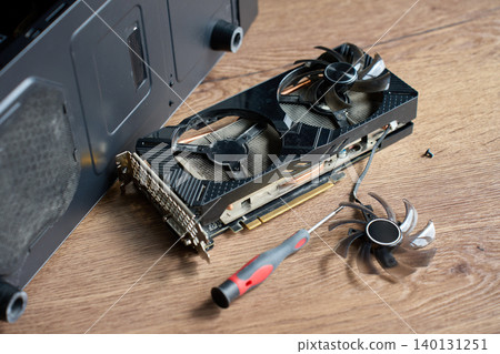 Disassembled graphics card, fan blade, screwdriver, and screw lie on wooden table near open PC case. Concept of computer repair, cleaning, and hardware maintenance. Disassembled graphics card, fan blade, screwdriver, and screw lie on wooden table near open PC case. Concept of computer repair, cleaning, and hardware maintenance. 140131251